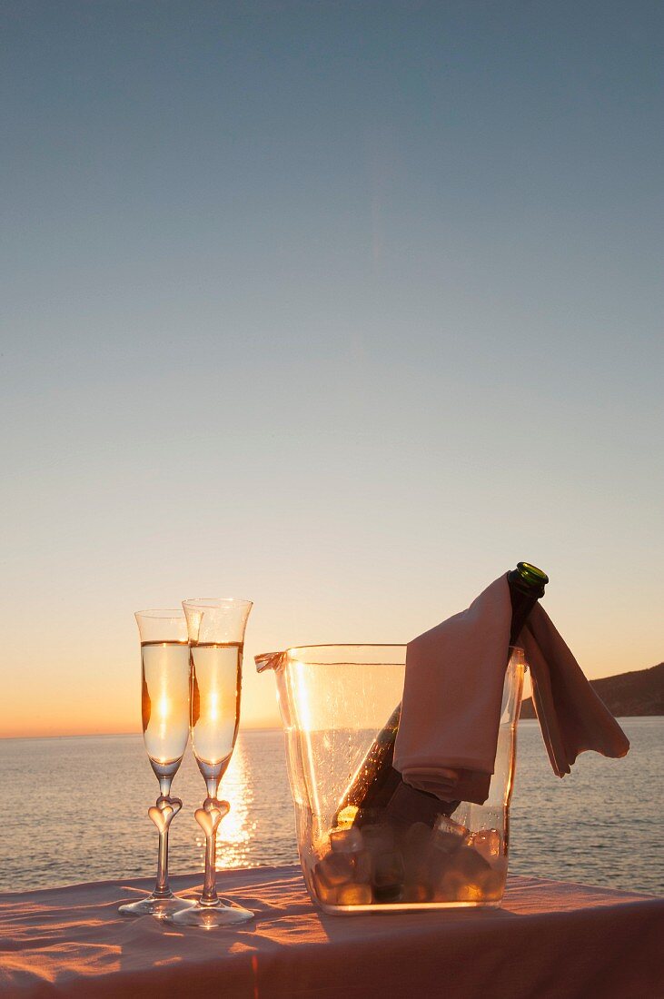A bottle of champagne in a cooler and two filled glasses against a sunset over the sea