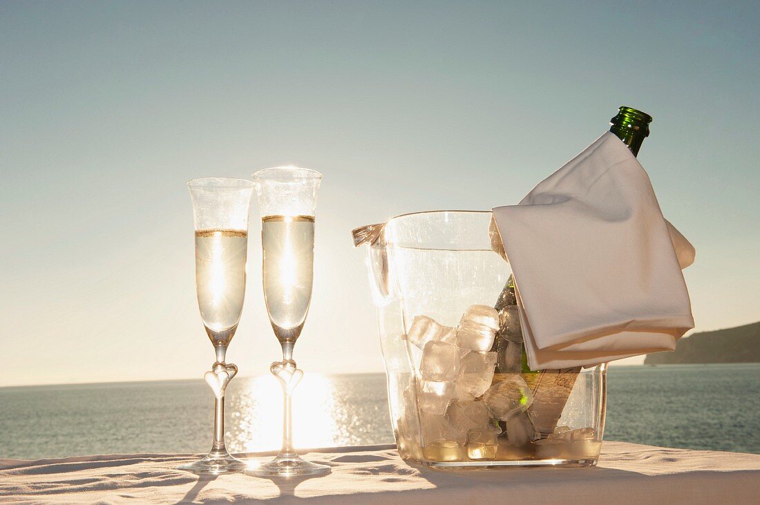 A bottle of champagne in a chiller and two glasses of champagne with a view of a sunset by the sea
