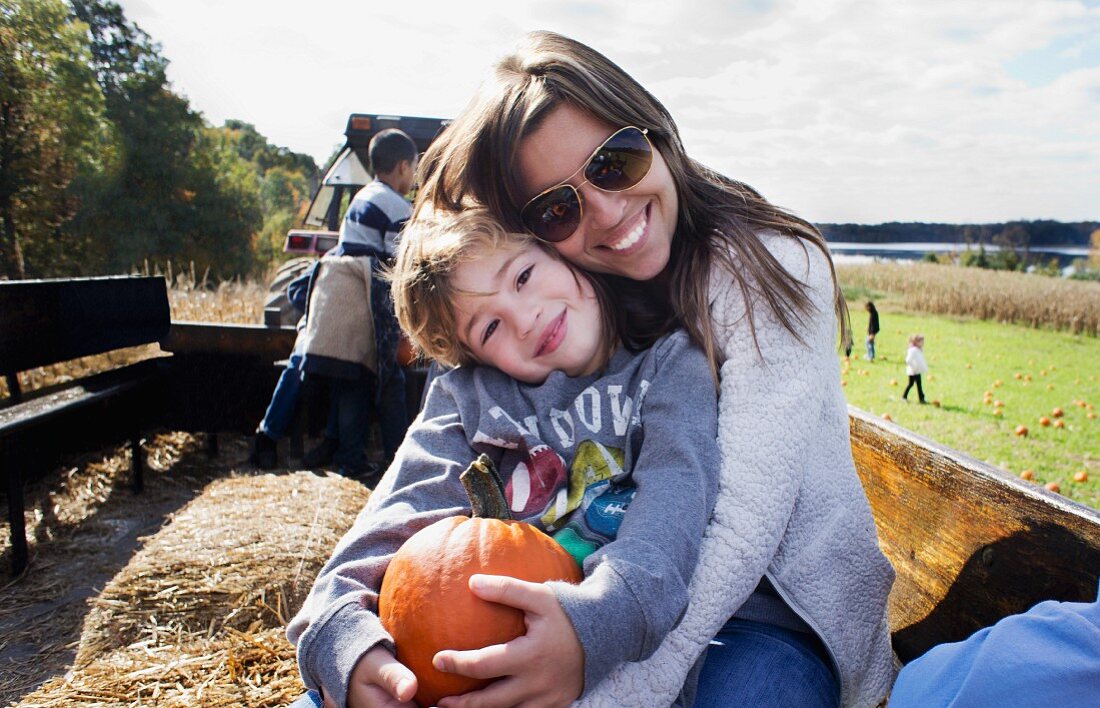 A mother and a son sitting in a tractor trailer holding a pumpkin