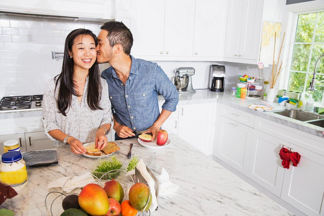 A man kissing a woman while preparing sandwiches in a kitchen