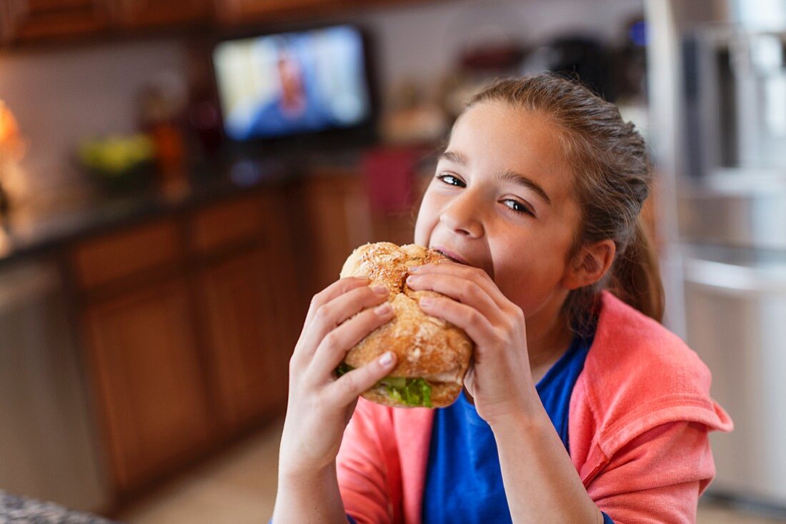 A girl in a kitchen biting into a large sandwich