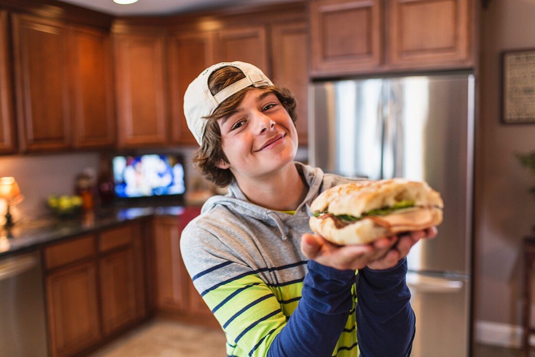 A teenage boy in a kitchen holding a large sandwich