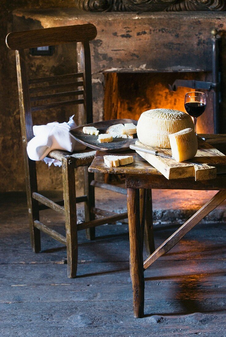 A wheel of pecorino on a chopping board with a glass of red wine in front of an open fireplace