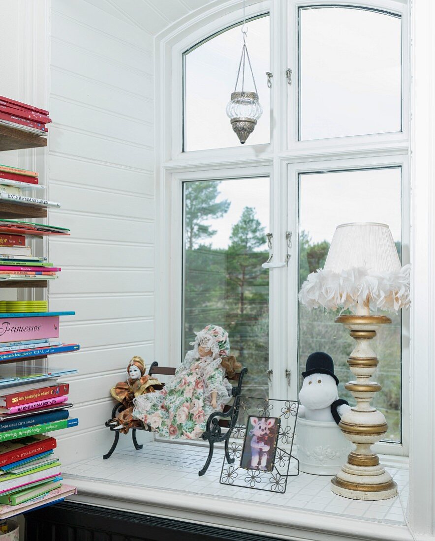 Puppen, Tierfigur, Tischleuchte und Fotorahmen in Fensternische; seitlich Bücherregal