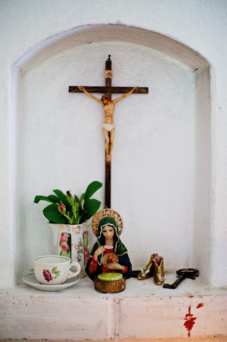 Kleine Wandnische mit Devotionalien, Heiligenfigur neben Tasse und Blumen in Krug, an Wand Kruzifix