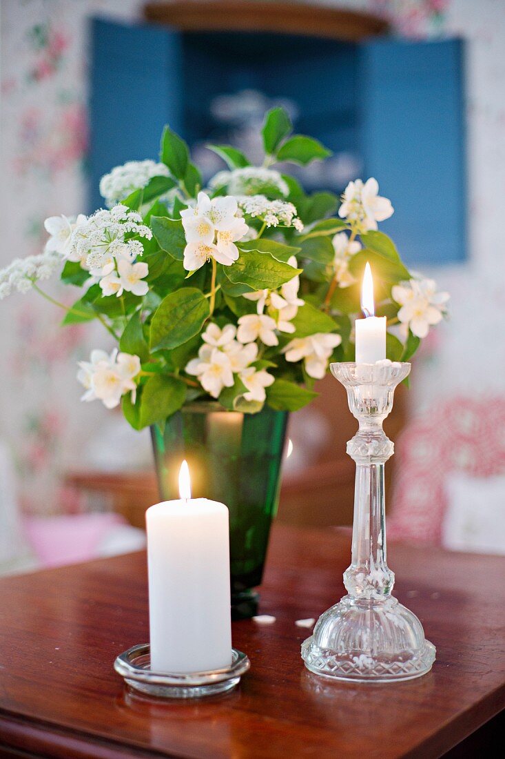 Pillar candle and standard candle in glass candle holders in front of green vase of jasmine on wooden table