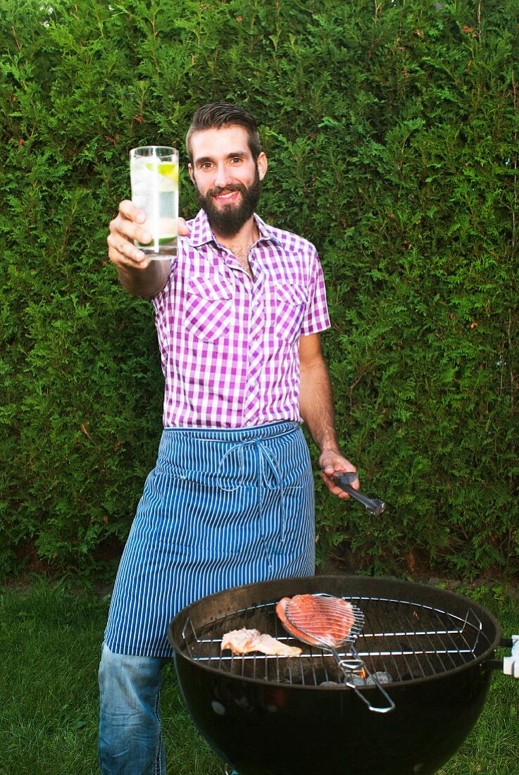 Junger Mann mit Schürze vor Grill mit Lachs und Forelle hält Drink