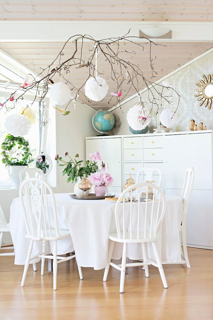Weiß lackierte Holzstühle um Tisch mit Tischdecke vor halbhohem Schrank, Zweig mit weissen Dekokugeln an Decke aufgehängt