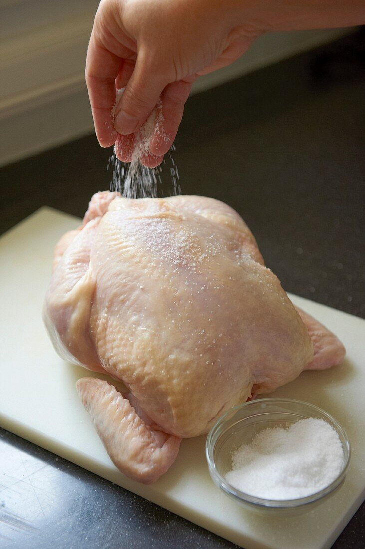 A raw chicken being seasoned with salt