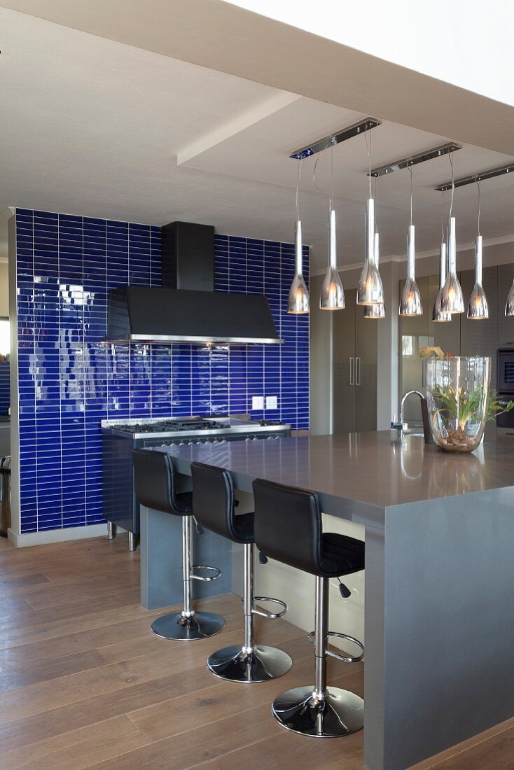 Bar stools with black leather covers at grey kitchen island below several rows of pendant lamps; wall of indigo tiles in background