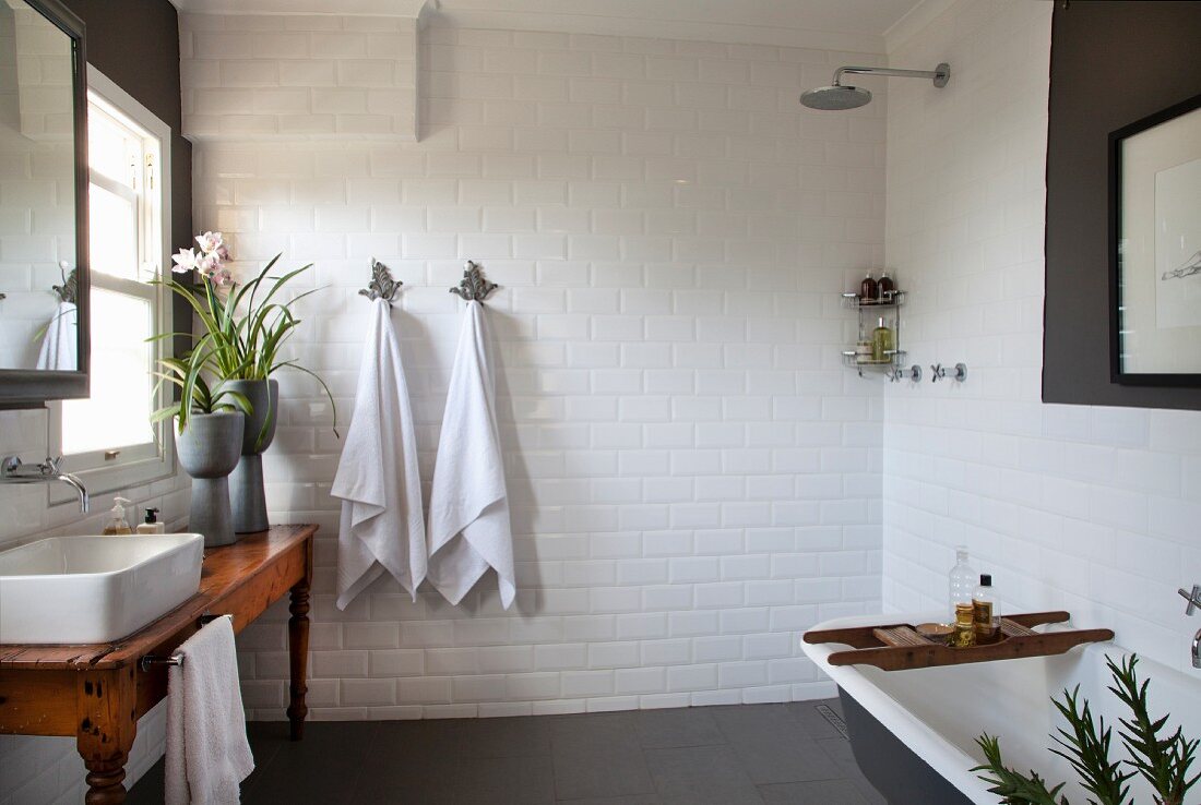 Rustic wooden table with countertop sink below window, open shower and bathtub in white-tiled bathroom