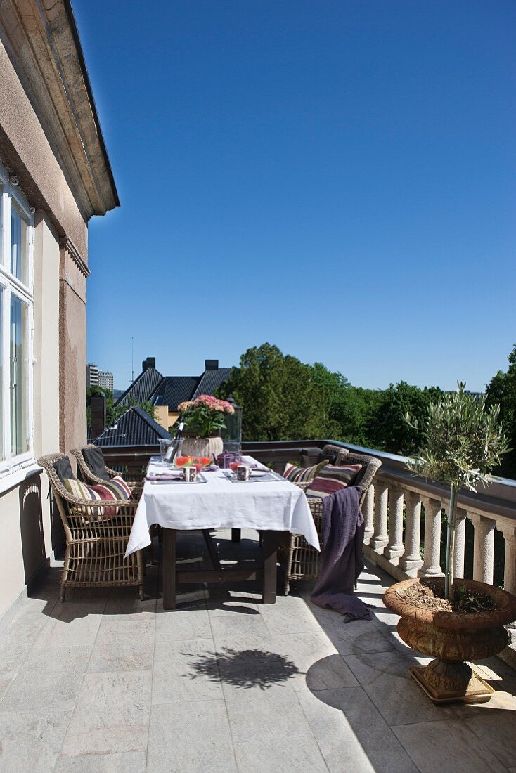 Rattanstühle um gedeckten Tisch mit weisser Tischdecke, auf sonnigem Balkon mit Steinbalustrade