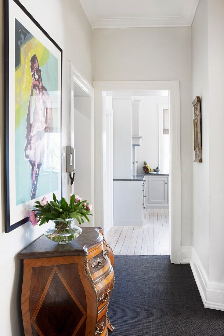 Rococo-style chest of drawers and modern artwork in hallway; open door leading to kitchen in background