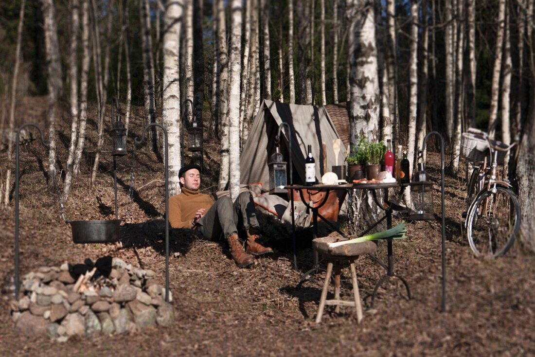 Feuerstelle und Picknickplatz mit Tisch, Zelt und Fahrrad im herbstlichen Wald, entspannter Mann am Baum sitzend