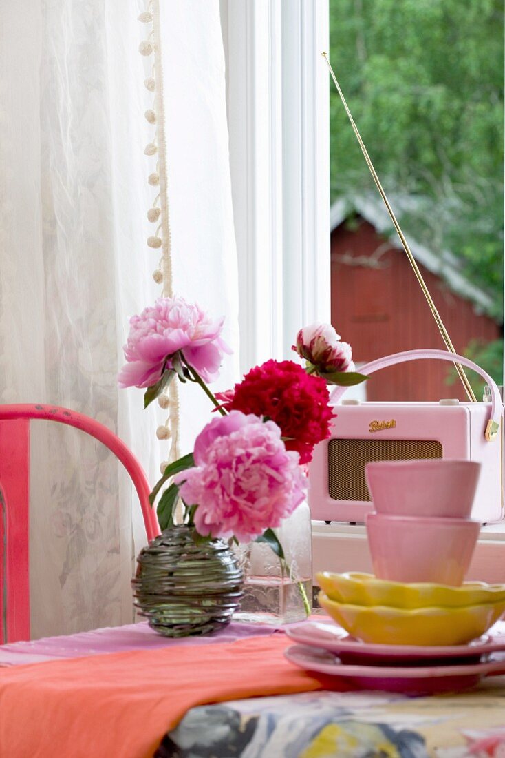 Vintage accessories in shades of pink and red with stacked crockery, retro radio and vase of peonies