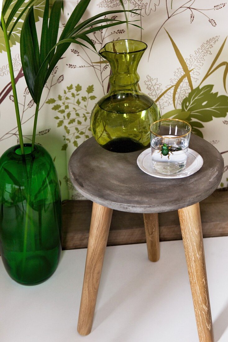 Vintage Holzhocker mit grüner Karaffe und Trinkglas, smaragdgrüne Bodenvase vor Tapete mit Blättermotiv