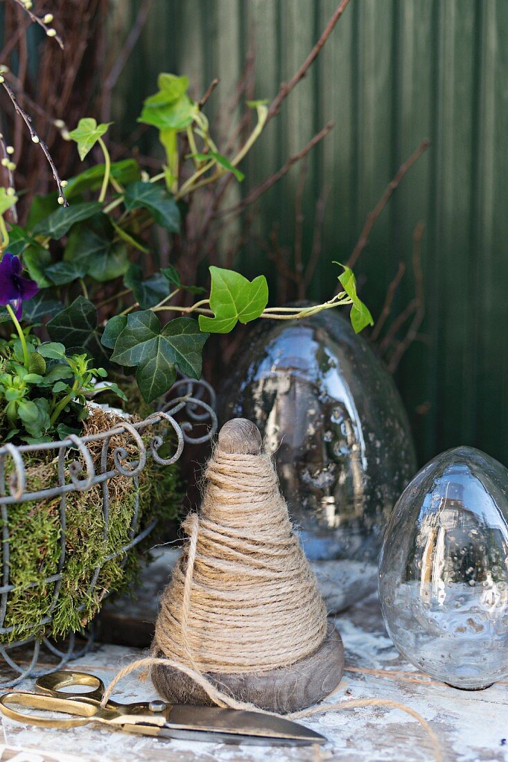 Wooden cone of twine, scissors and glass eggs next to Easter arrangement in wire basket