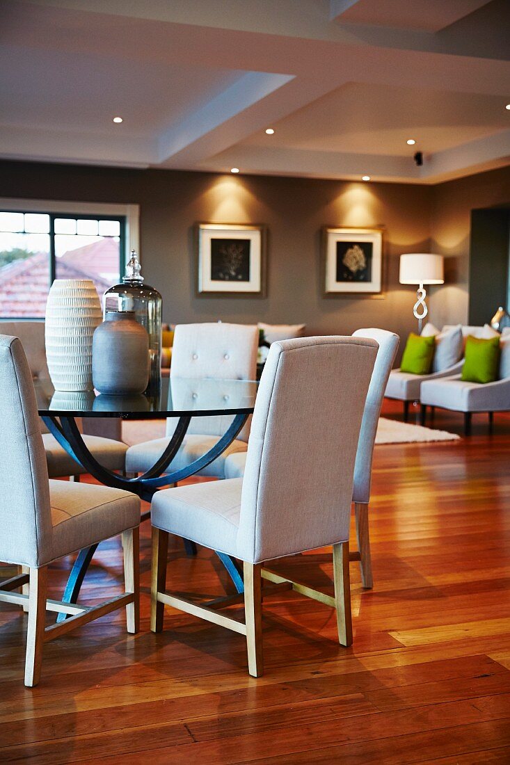 Various vases on round glass table and white upholstered chairs; easy chairs under ceiling spotlights in corner of grey-painted room