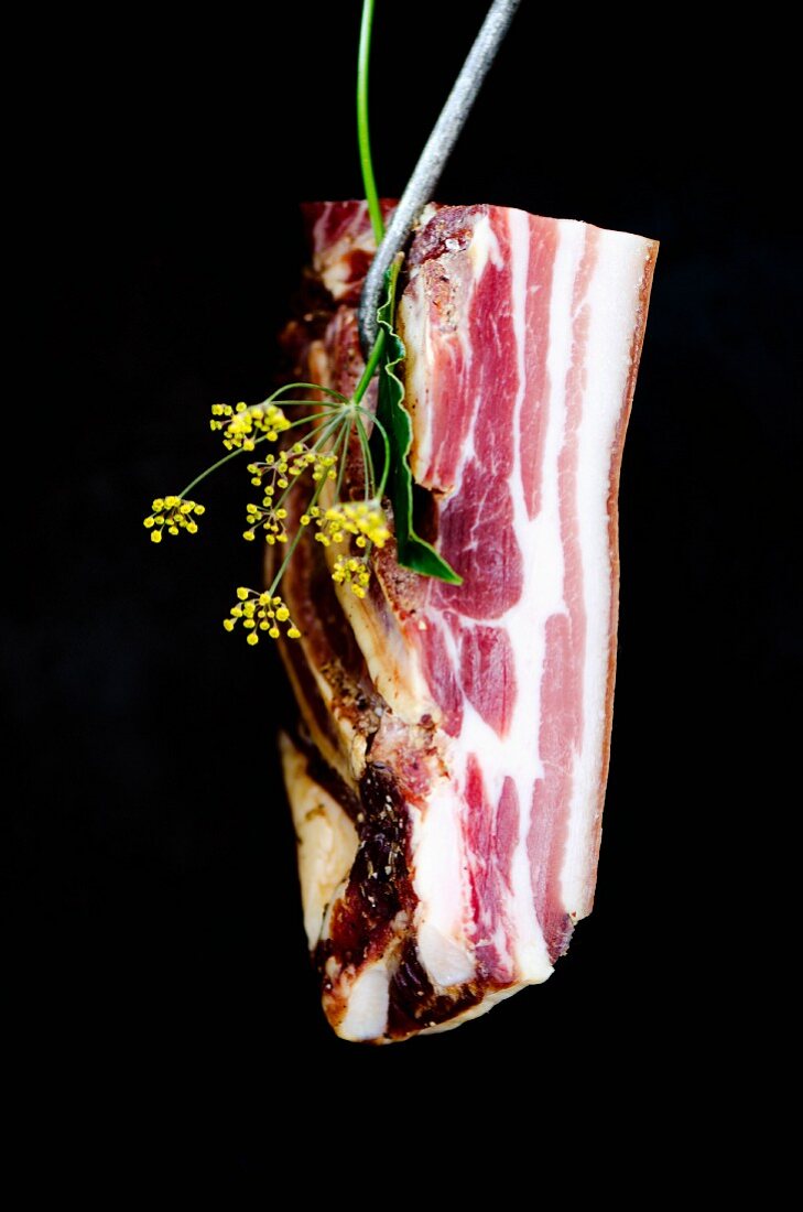 Pancetta hängt an einem Fleischerhaken mit frischen italienischen Kräutern