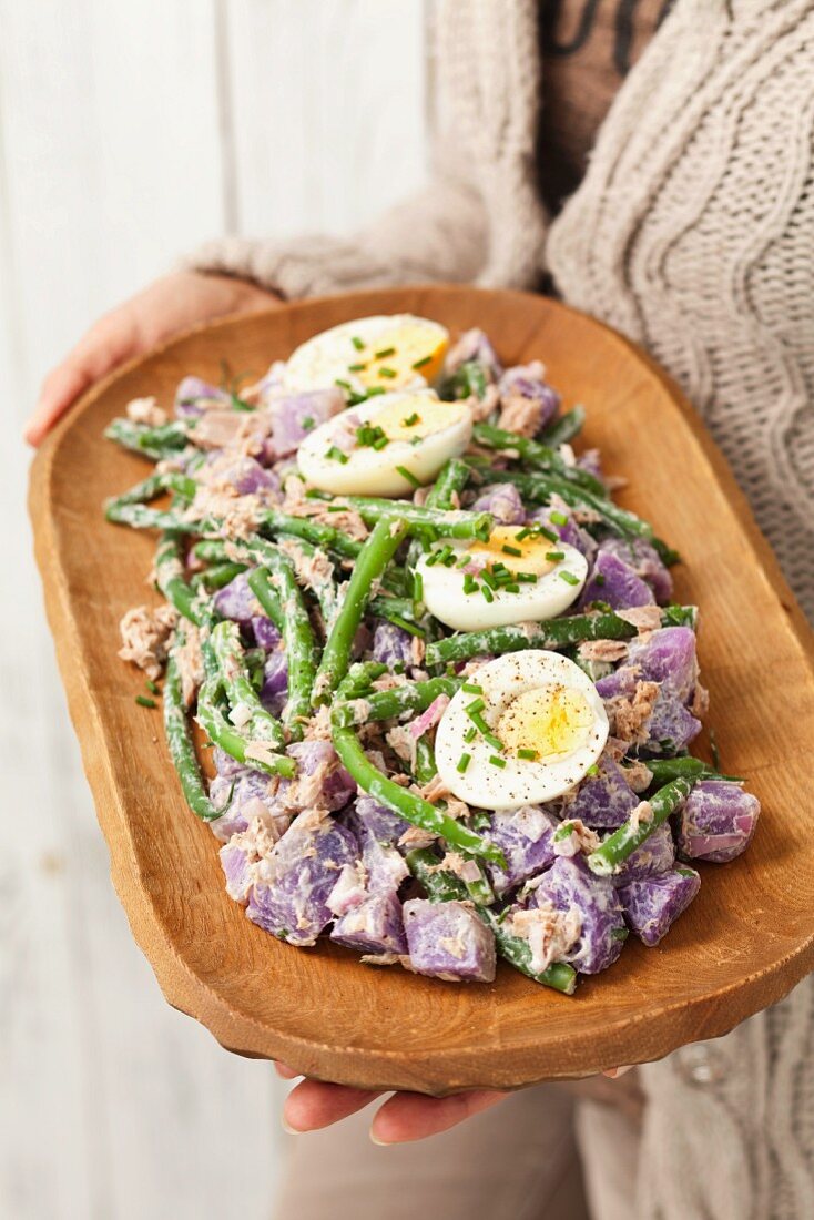 Frau hält Holzschale mit lila Kartoffelsalat mit grünen Bohnen, Ei, Thunfisch und Mayonnaise