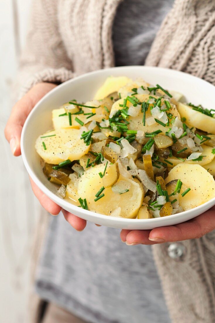 A woman holding a bowl of potato salad with gherkins, onions and chives