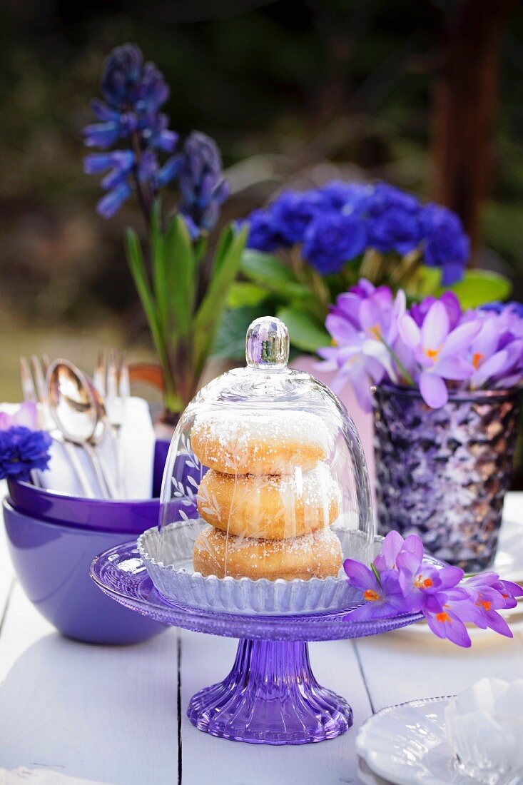 Doughnuts under a glass cloche on a table outside decorated with crockery and blue and purple flowers