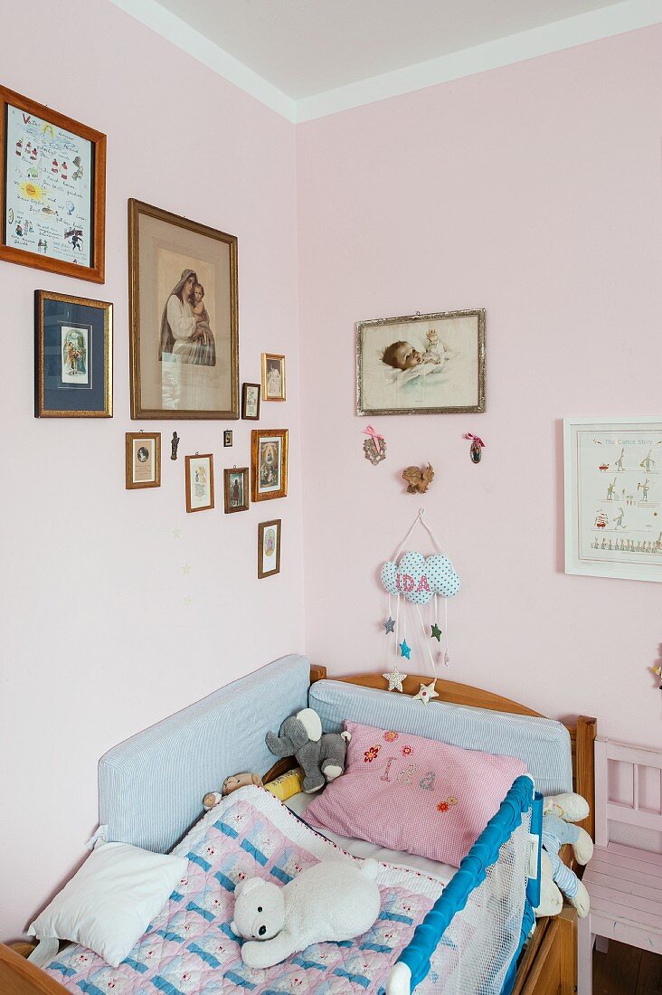 Soft toys on child's bed in corner below framed pictures on pink-painted walls