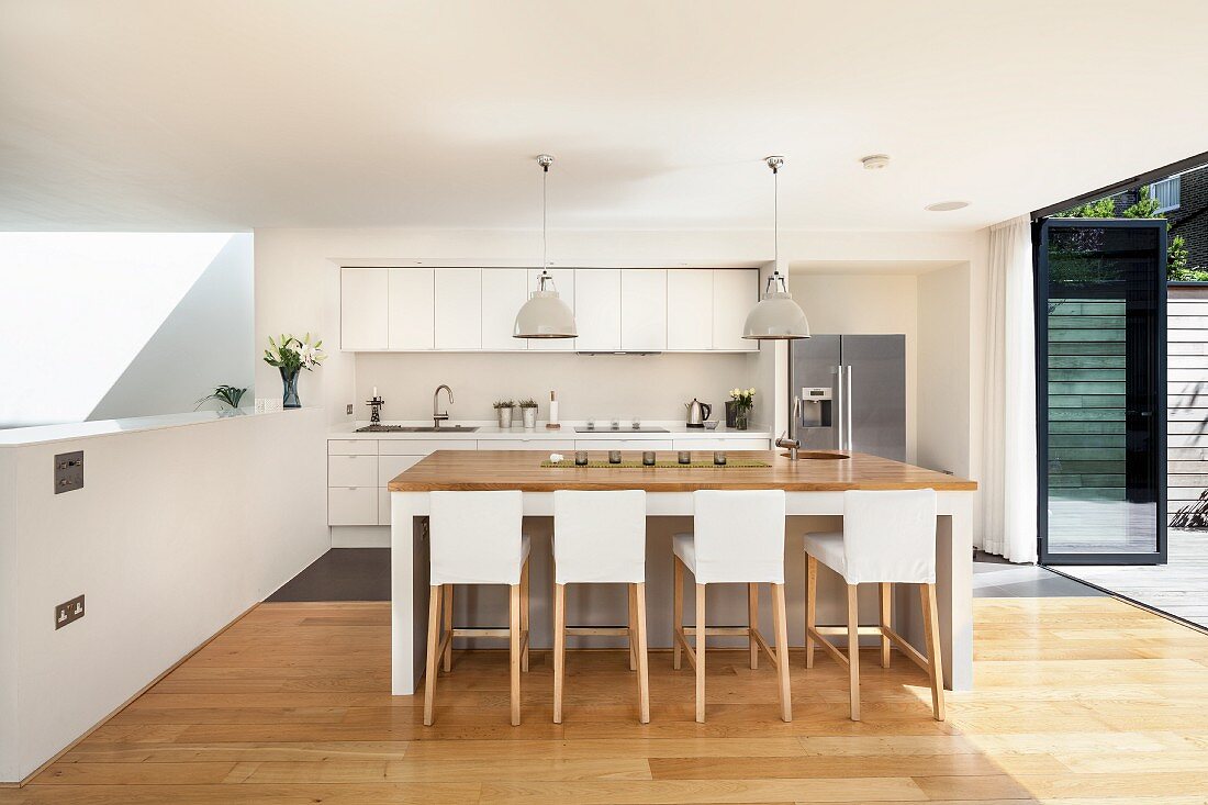 Bar stools with white loose covers in open-plan designer kitchen with open folding terrace doors to one side