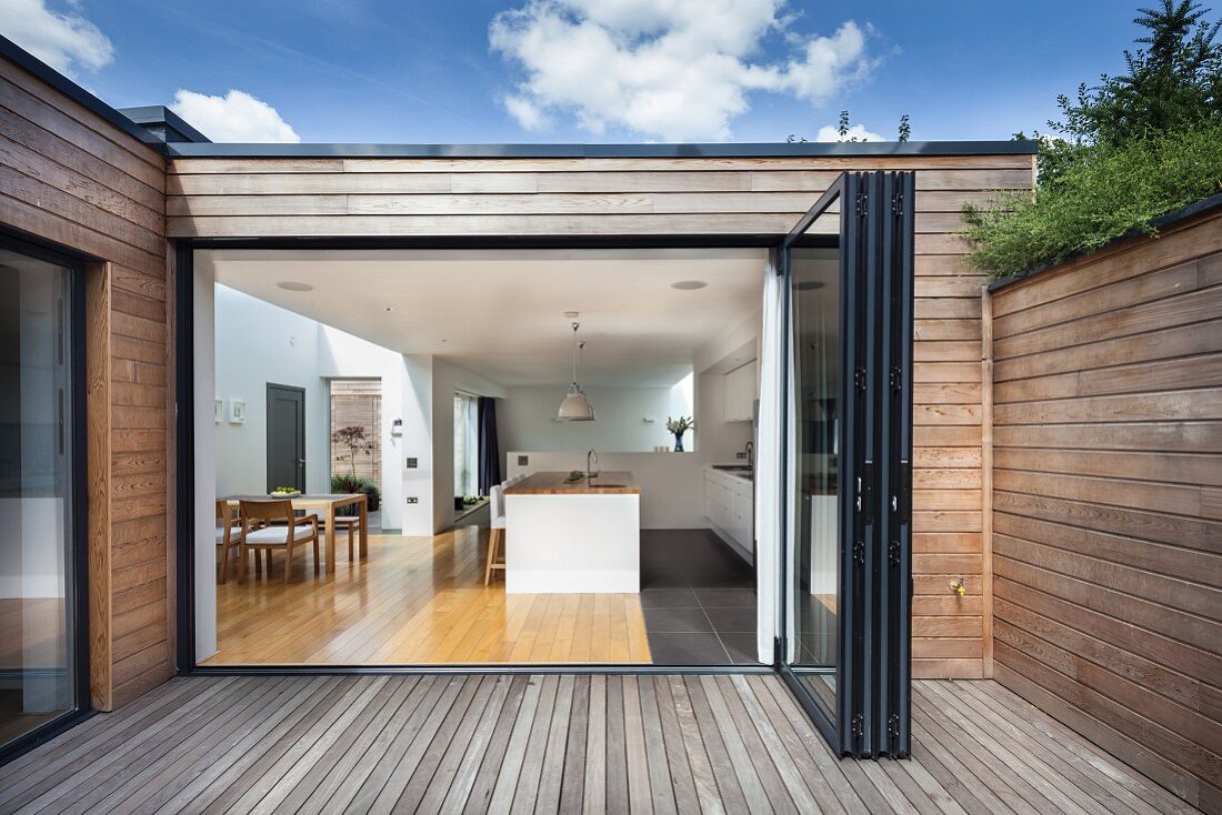 Wooden terrace of contemporary house with wood-clad facade; view through open folding door into kitchen