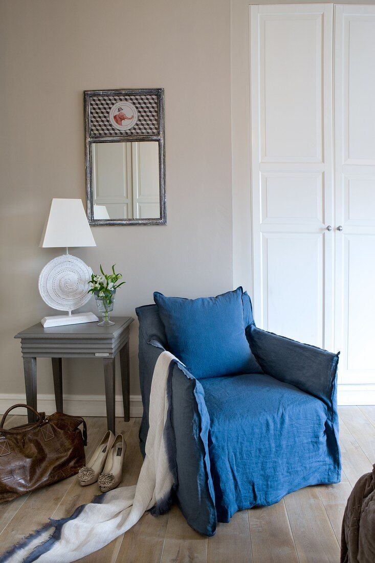 Lady's accessories next to blue, loose-covered armchair in bedroom next to lamp on table below mirror