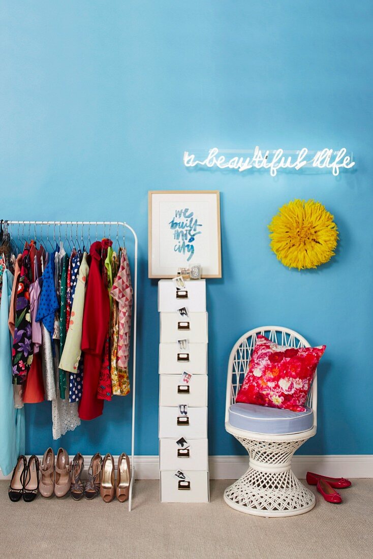 Clothes hanging on rail next to stack of white storage boxes and wicker chair under illuminated neon lettering on wall painted light blue