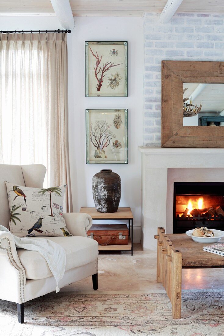 Country-style living room with wing-back chair and coffee table in front of open fire and whitewashed brick wall