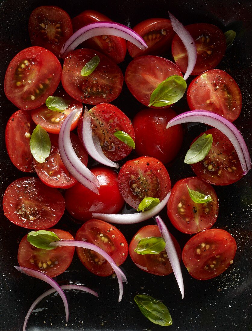 Halved plum tomatoes with red onion slices and basil leaves in a roasting tin