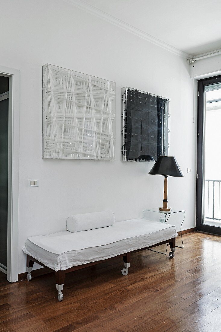 Couch on castors with white loose cover next to table lamp on transparent side table below black and white artworks on wall