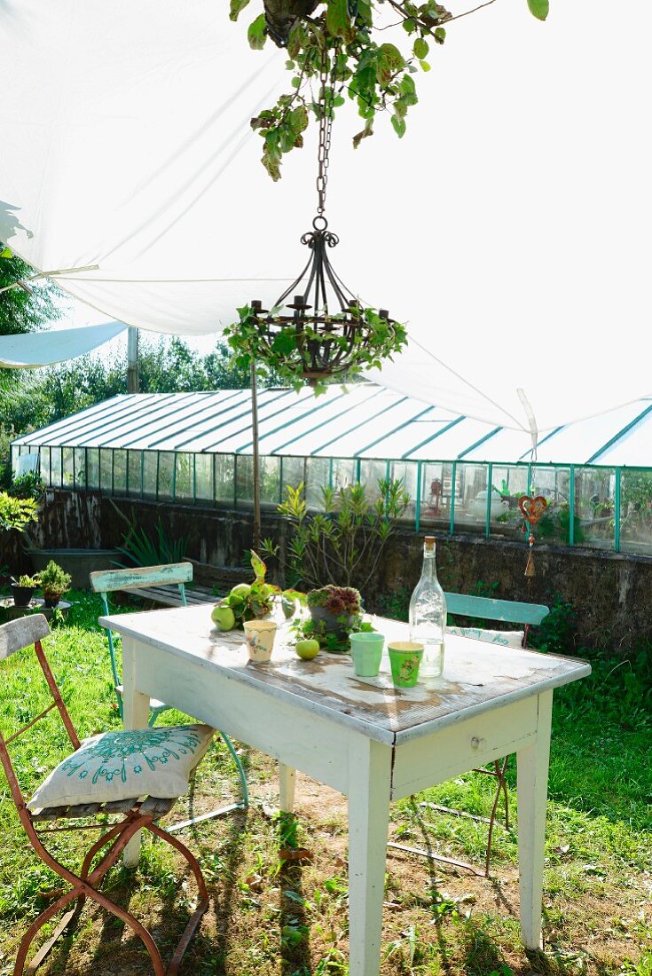 Old garden chairs with cushions at rustic garden table with beakers and vintage lemonade bottle below chandelier wreathed in ivy and awning; greenhouse in background