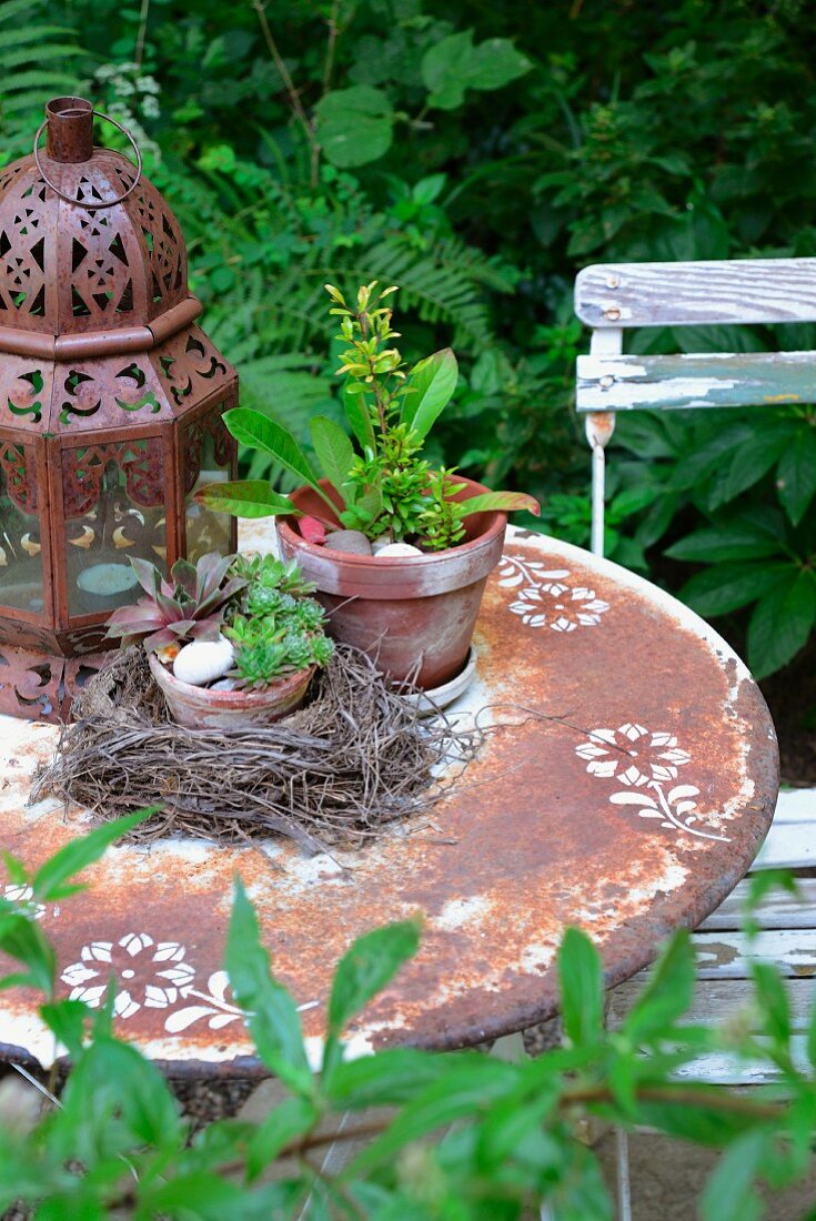 Alter Gartentisch mit Rost-Patina und schabloniertem Blumendekor, darauf orientalische Laterne, Vogelnest mit Hauswurz und Pflänzchen im Tontopf