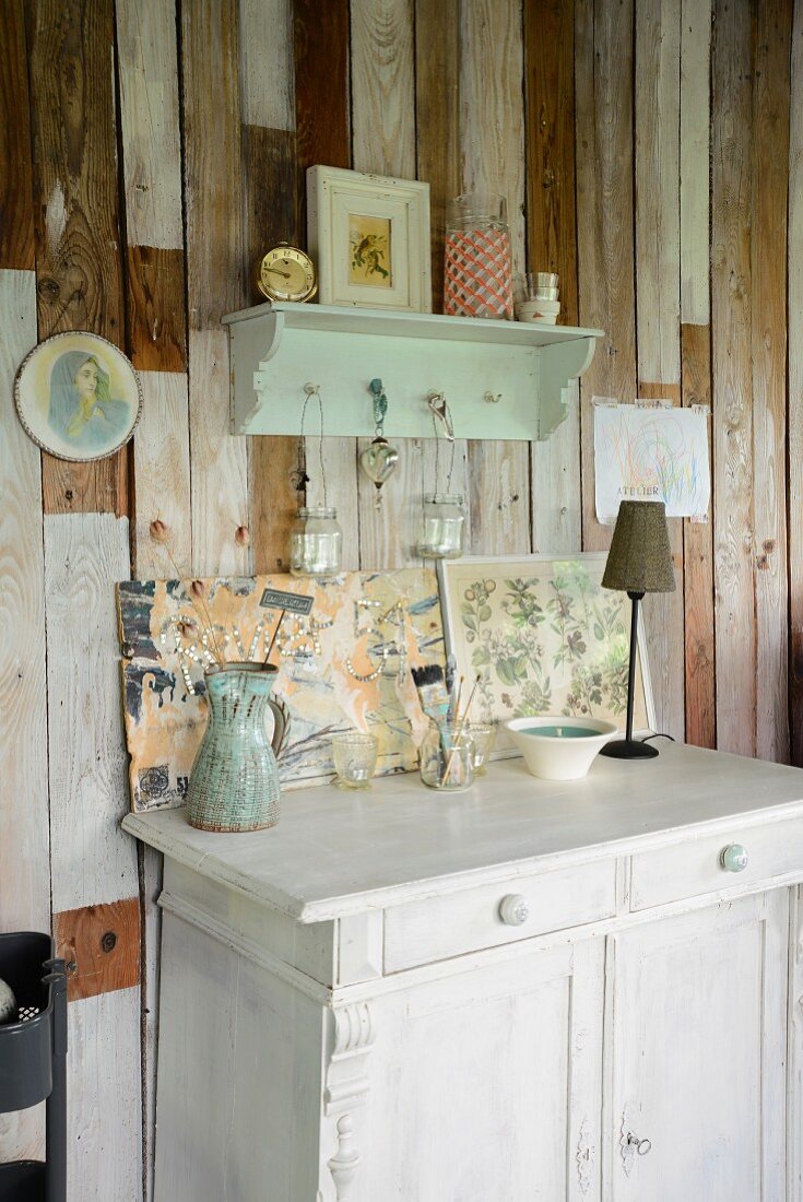 Jug, paintbrushes in glass jar and table lamp on old, white cabinet below small wall bracket on simple board wall