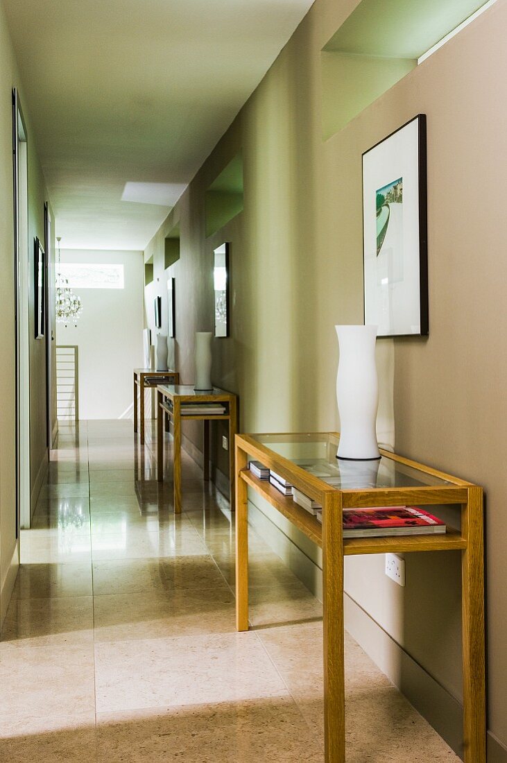 Three white vases on three wooden console tables with glass tops below framed pictures on sand-coloured wall in long, narrow hallway