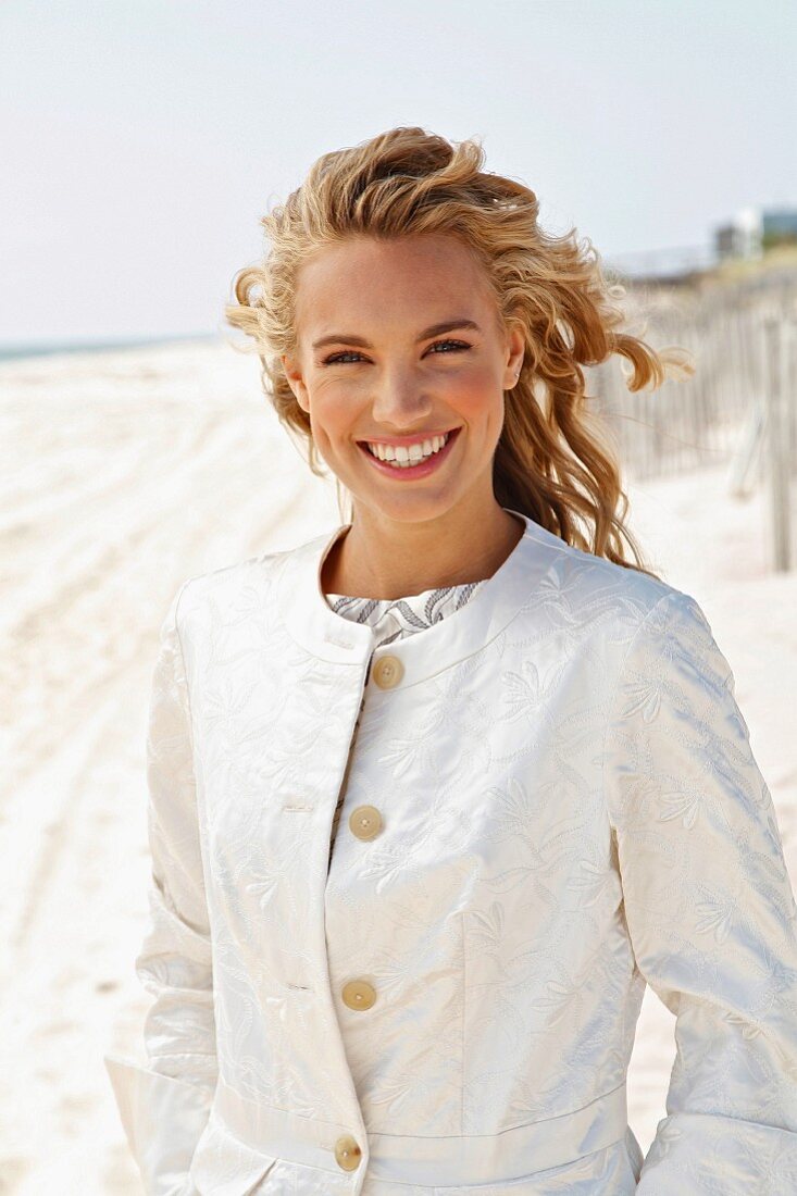 A young blonde woman on a beach wearing a white coat