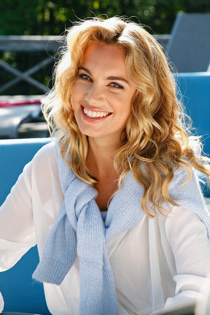 A young blonde woman outside wearing a white blouse with a jumper over her shoulders