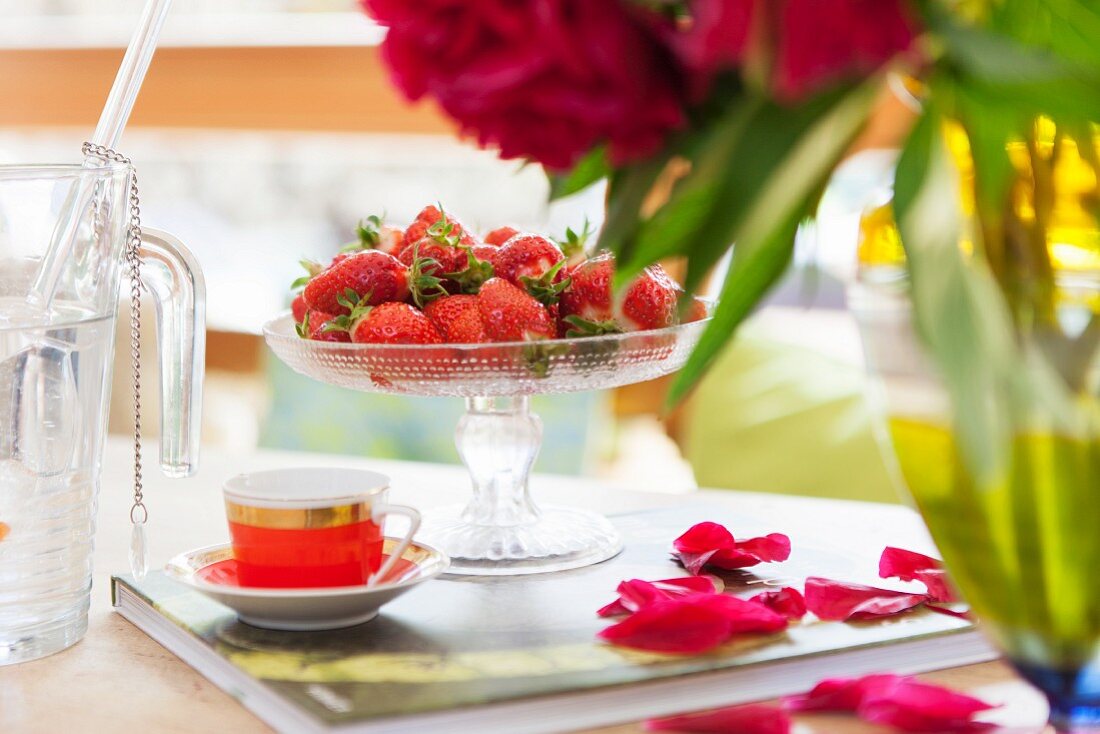 Frische Erdbeeren auf Glasschale und Mokkatasse neben verstreuten Blütenblättern auf Buch