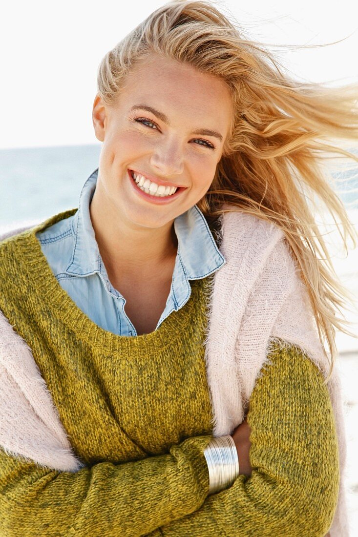 A young blonde woman by the sea wearing a knitted jumper and a denim shirt with another jumper over her shoulders