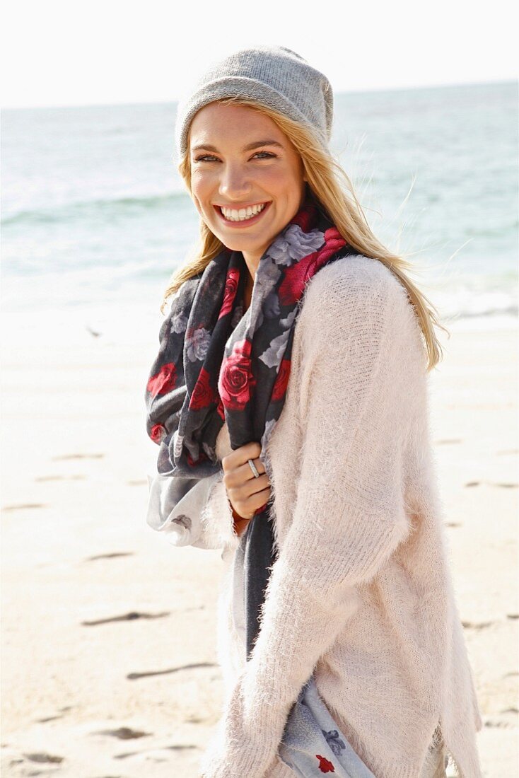 A young blonde woman by the sea wearing a light jumper, a scarf and a hat