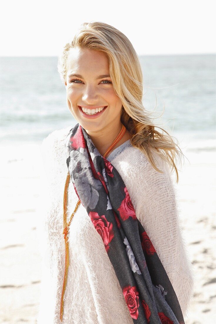 A young blonde woman by the sea wearing a light jumper and a scarf