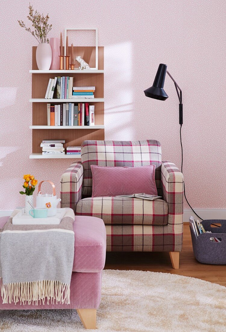 Gemütlicher Sessel mit Karomuster und violettem Kissen, gegenüber gepolstertem Hocker, schwarze Wandleuchte und Regal Board an pastellrosa getönter Wand