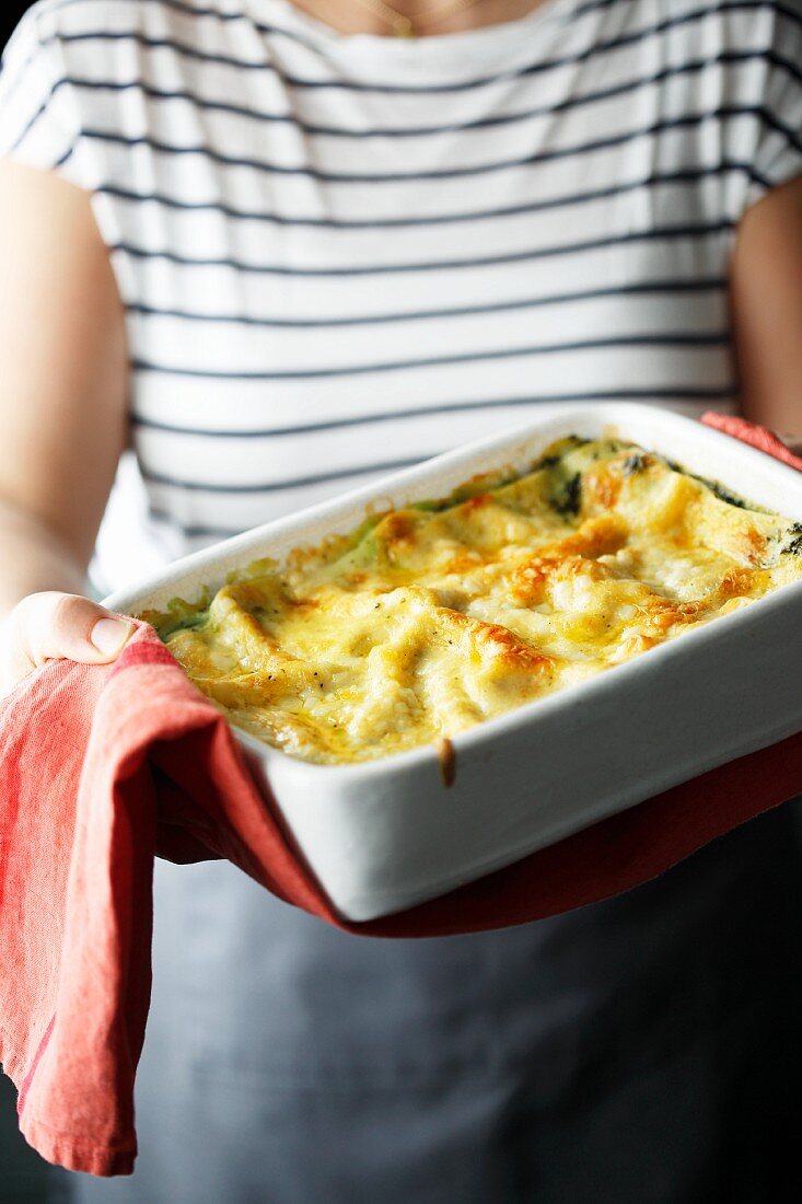 A woman holding a baking tray of spinach lasagne with chilli
