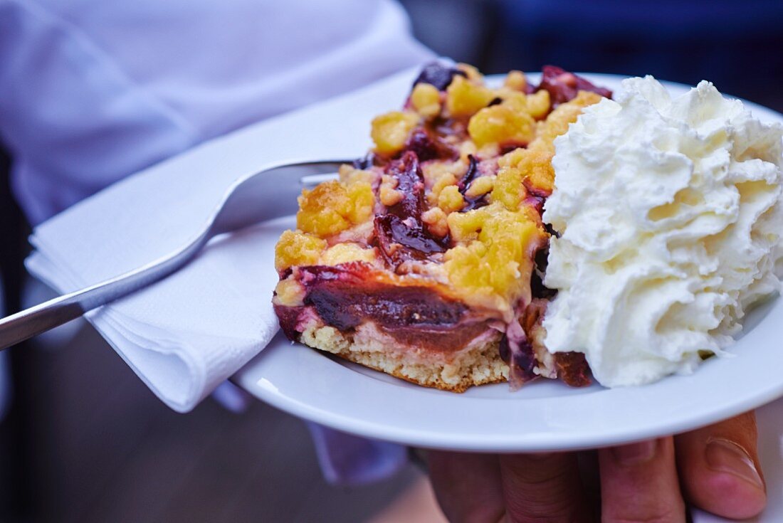 Koch hält Teller mit einem Stück Pflaumen-Streusel-Kuchen und Schlagsahne