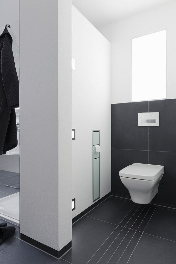 Raumteiler zwischen Dusche und Toilette mit integriertem Papierspender und Einbauleuchten, auf Boden und teilweise an Wand schwarze, grossformatige Fliesen