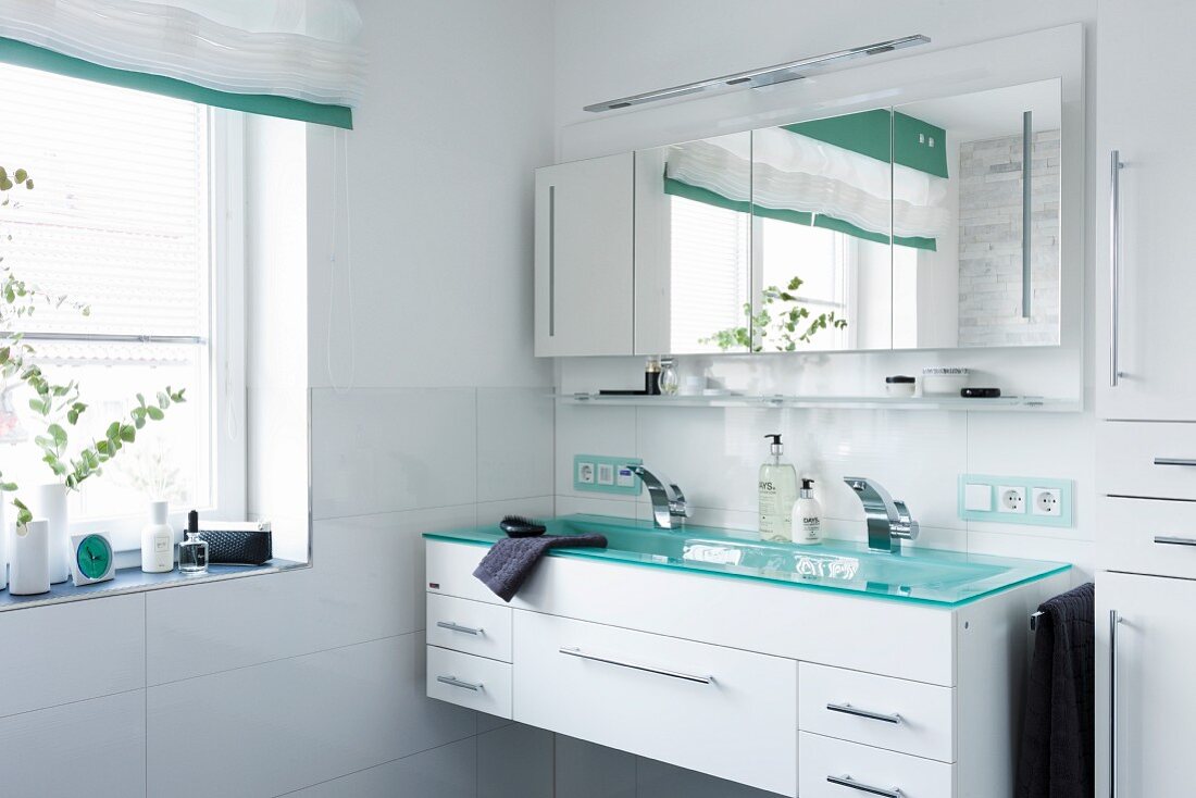 Trogartiger Waschtisch für Zwei mit türkis schimmernder Glasplatte auf weißem Schrankunterbau, oberhalb viertüriger Spiegelschrank, in modernem Bad mit Fenster