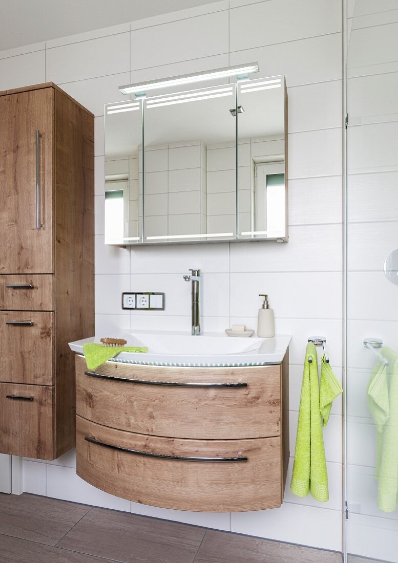 A curved, oak-style washstand on a white tiled wall with a three-section mirrored cabinet above it with an integrated strip light